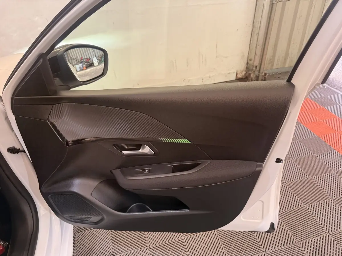 Vue côté gauche de la porte avant blanche d'une Peugeot 208 2025, avec garniture noire et insert effet carbone.
