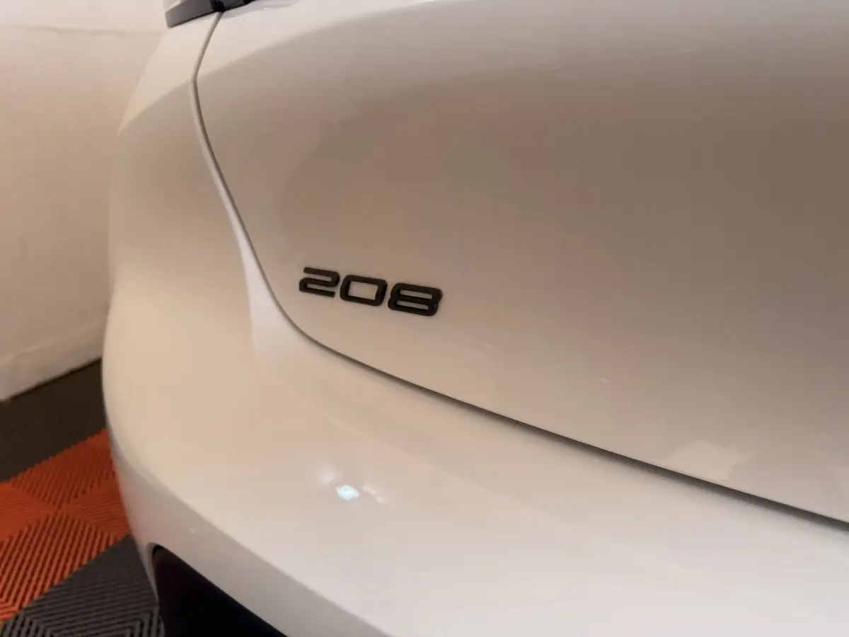 Gros plan sur le logo 208 à l'arrière gauche d'une Peugeot 208 blanche, avec sol en damier noir et orange.