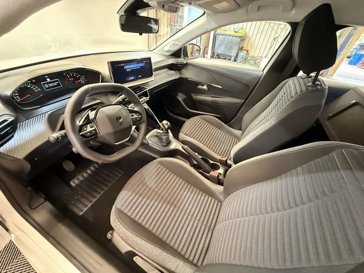 Vue intérieure avant droite de la Peugeot 208 blanche, montrant le volant cuir, tableau de bord digital et sièges gris.
