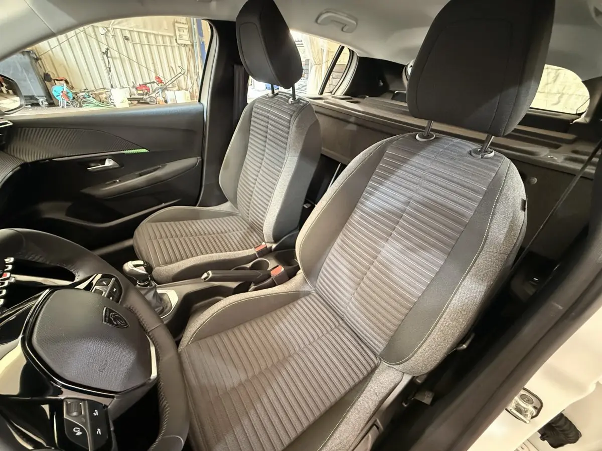 Vue intérieure côté conducteur de la Peugeot 208 blanche 2025, sièges tissu gris et volant cuir multifonctions.