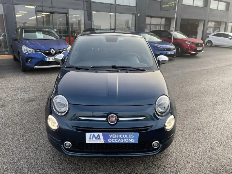 Vue avant d'une FIAT 500 bleu métal 2023 avec phares allumés, garée devant un showroom de véhicules d'occasion.