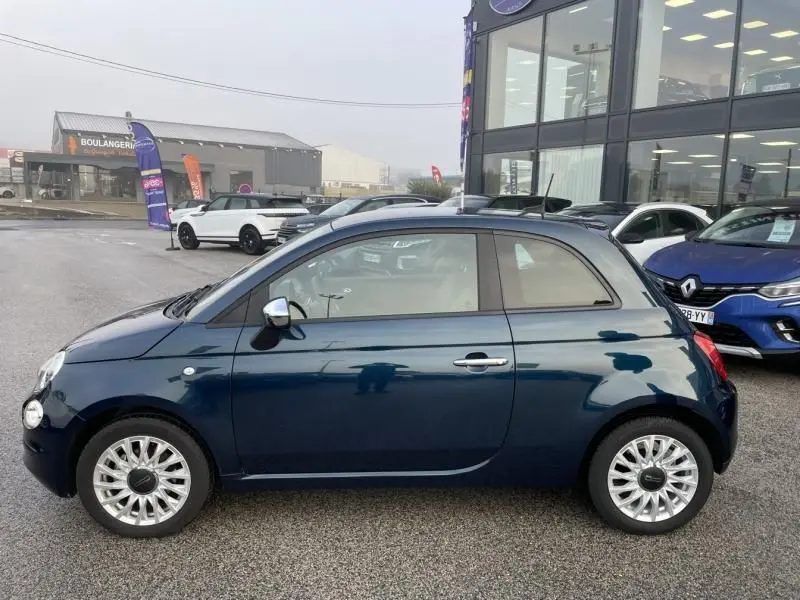 Vue de profil côté gauche d'une FIAT 500 bleu métal 2023 avec jantes alliage et rétroviseurs chromés, stationnée en extérieur.