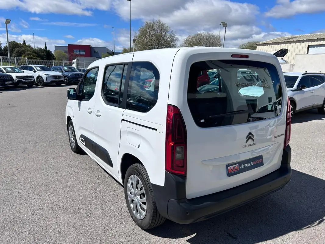 Vue 3/4 arrière droite d'un Citroën Berlingo blanc avec barres de toit et vitres teintées, stationné en extérieur.