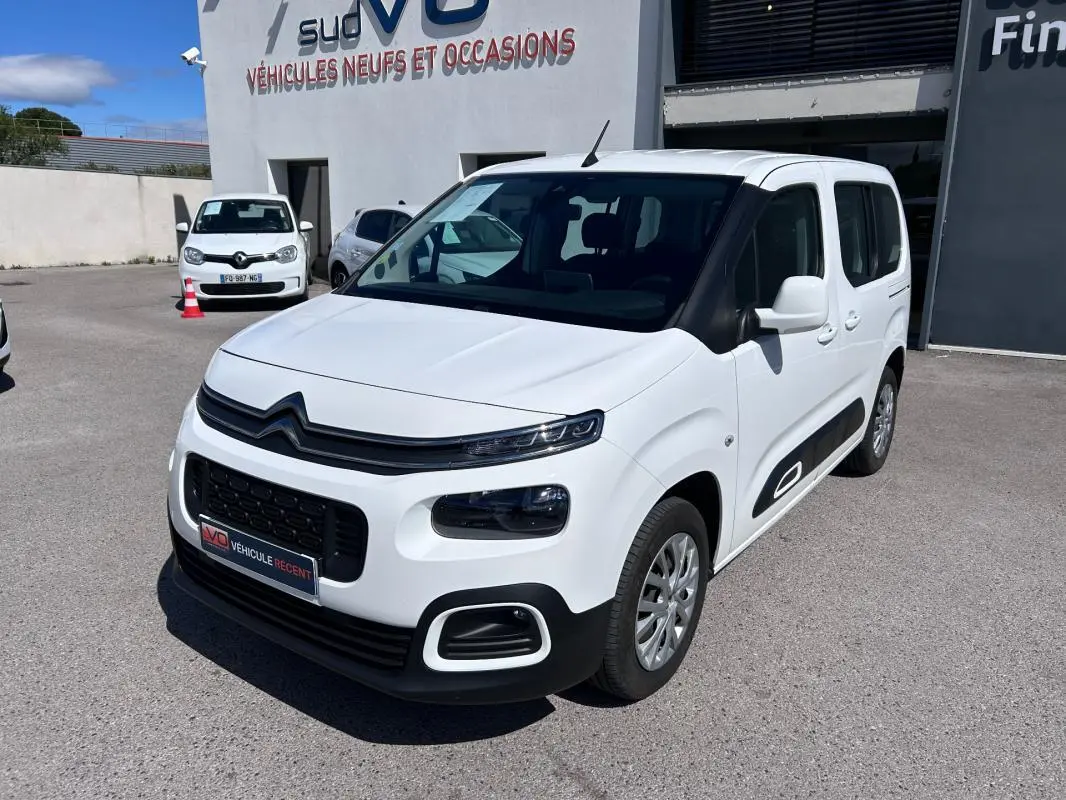 Citroën Berlingo blanc en 3/4 avant droit, avec barres de toit et protections latérales noires visibles.