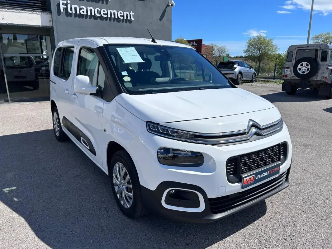 Vue 3/4 avant droite d'un Citroën Berlingo blanc avec barres de toit et protections latérales noires.