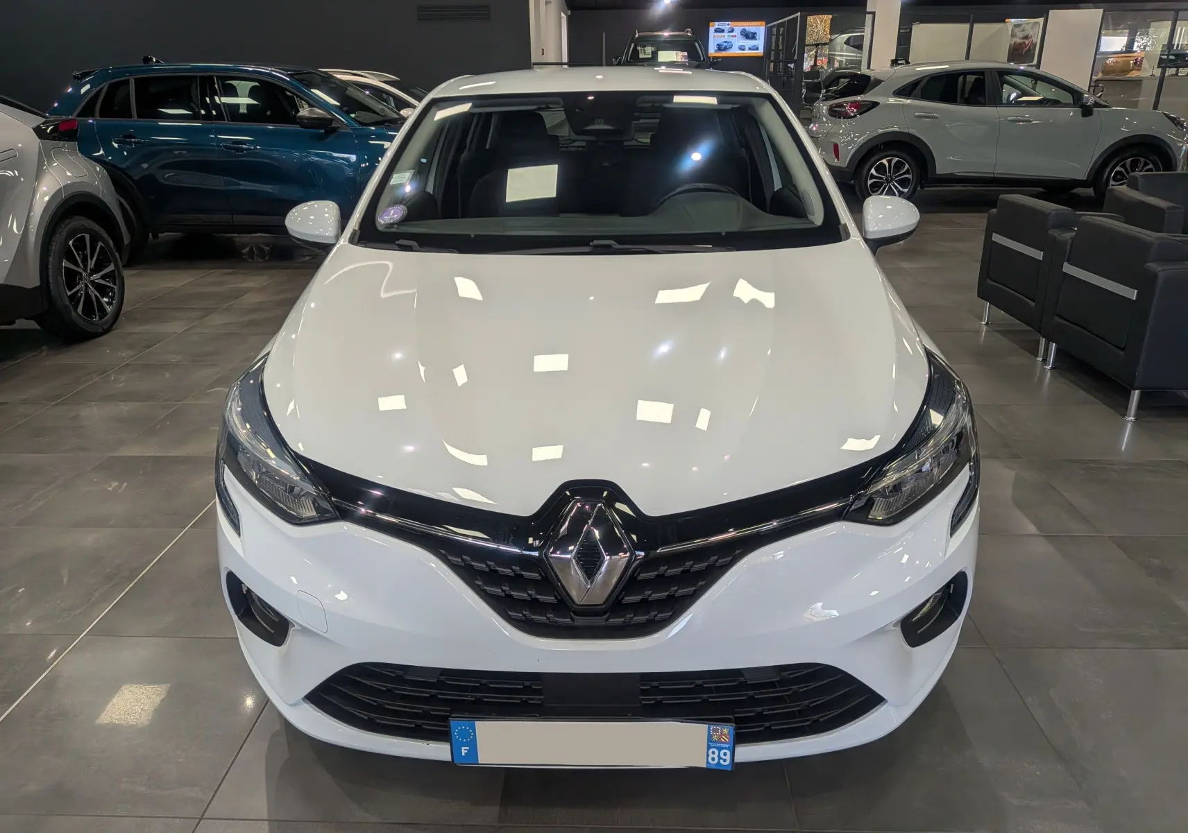 Vue de face d'une Renault Clio V TCe 100 Business en Blanc Glacier, éclairage showroom soulignant sa calandre noire brillante.