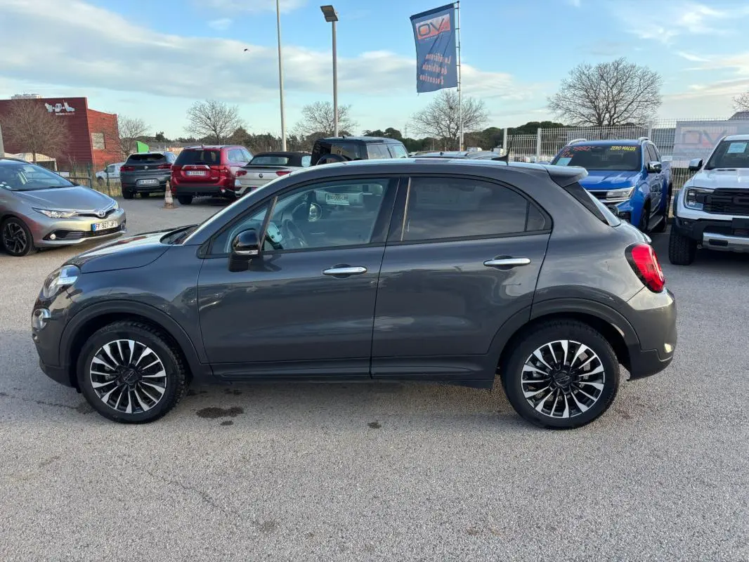 Profil droit d'un FIAT 500X gris 2023 avec jantes noires et argentées, stationné en extérieur sur un parking.