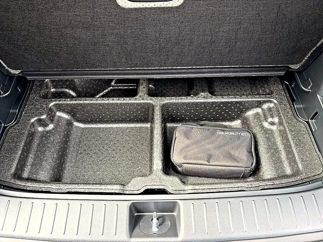 Coffre ouvert du Kia Sportage 1.6 hybride gris eclipse, vue sur le rangement sous plancher avec kit de mobilité pneus.
