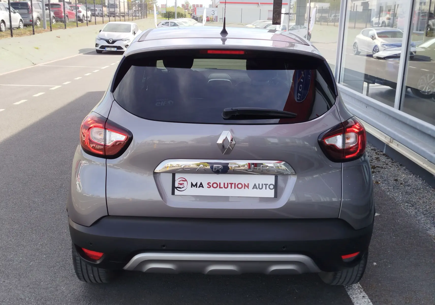 Vue arrière d'un Renault Captur gris Cassiopée 2018 avec feux arrière allumés et logo visible sur un parking.