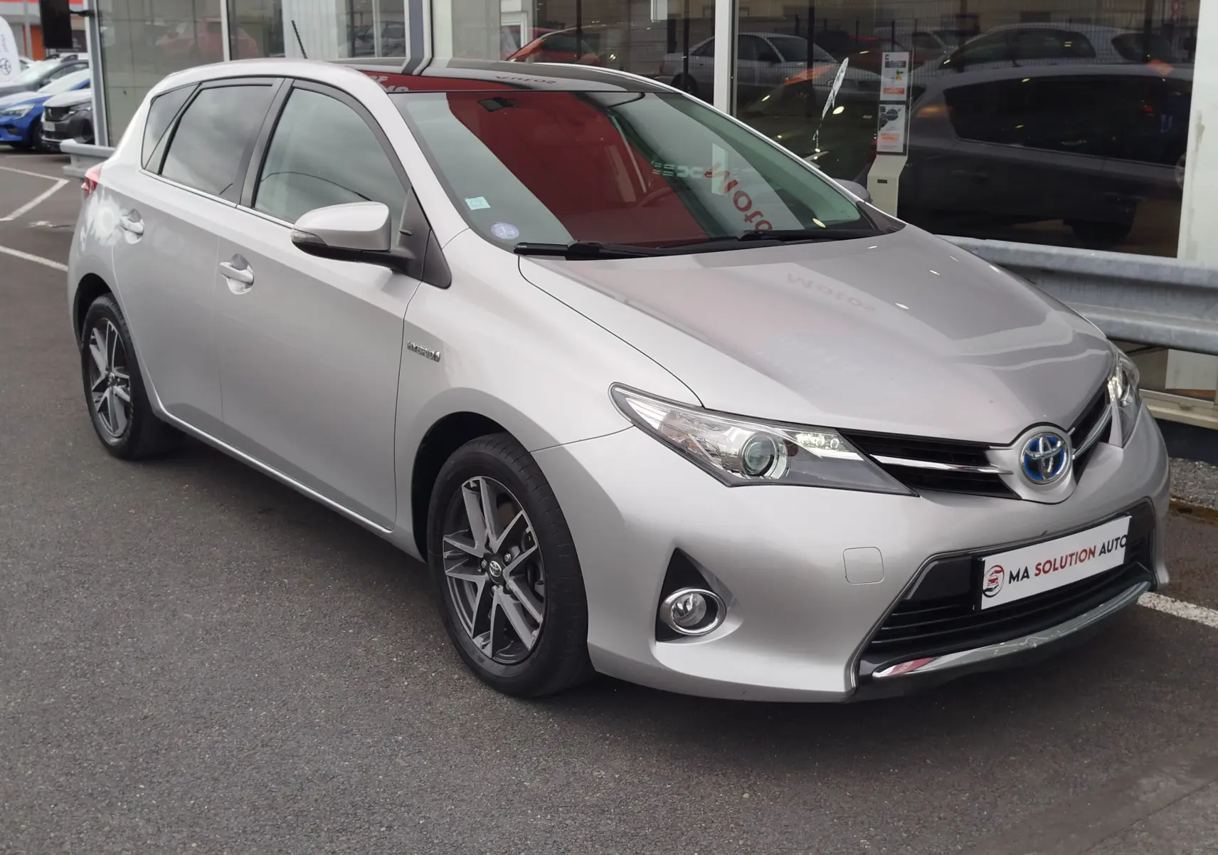 Toyota Auris HSD 136h gris aluminium vue 3/4 avant droit, avec jantes alliage et calandre distinctive hybride.