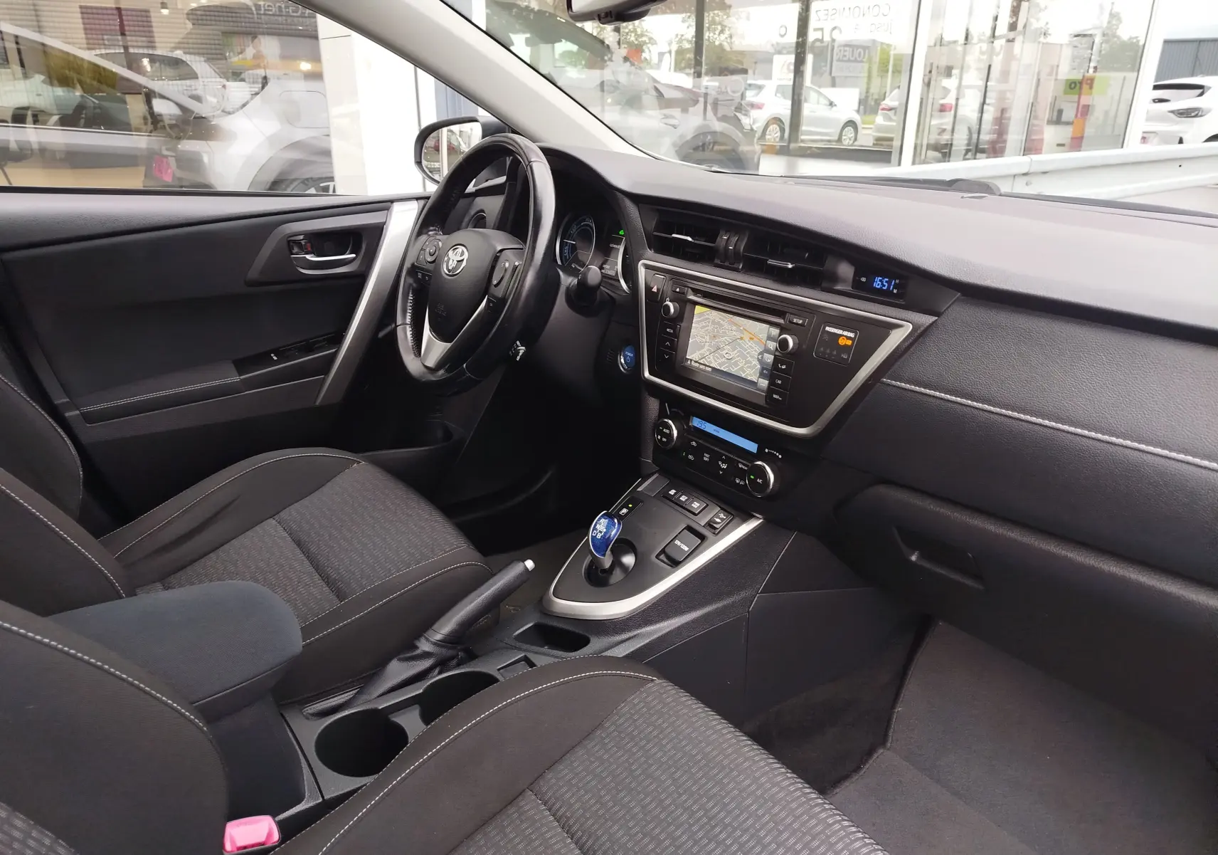 Vue intérieure côté conducteur de la Toyota Auris HSD 136h gris aluminium, avec tableau de bord et console centrale visibles.