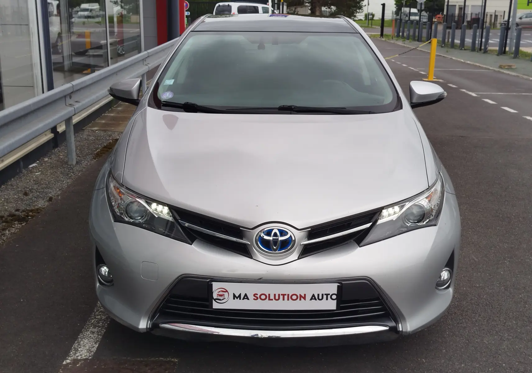 Vue avant d'une Toyota Auris HSD 136h gris aluminium, avec feux LED allumés et calandre noire brillante.