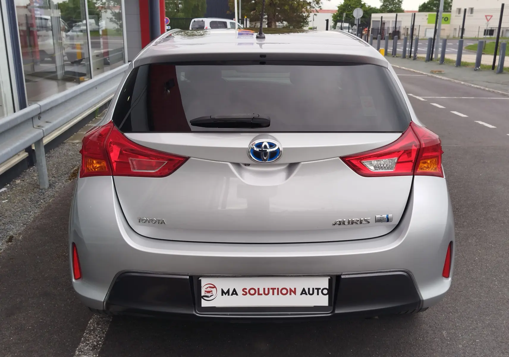 Vue arrière d'une Toyota Auris HSD 136h gris aluminium de 2015, avec feux arrière rouges et logo hybride visible.
