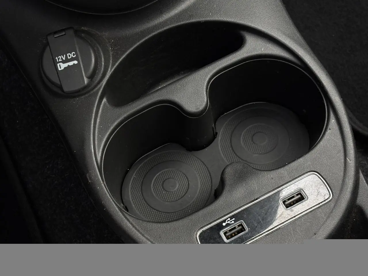 Gros plan sur la console centrale noire du Fiat 500 Hybrid 2024, avec porte-gobelets, prise 12V et ports USB visibles.