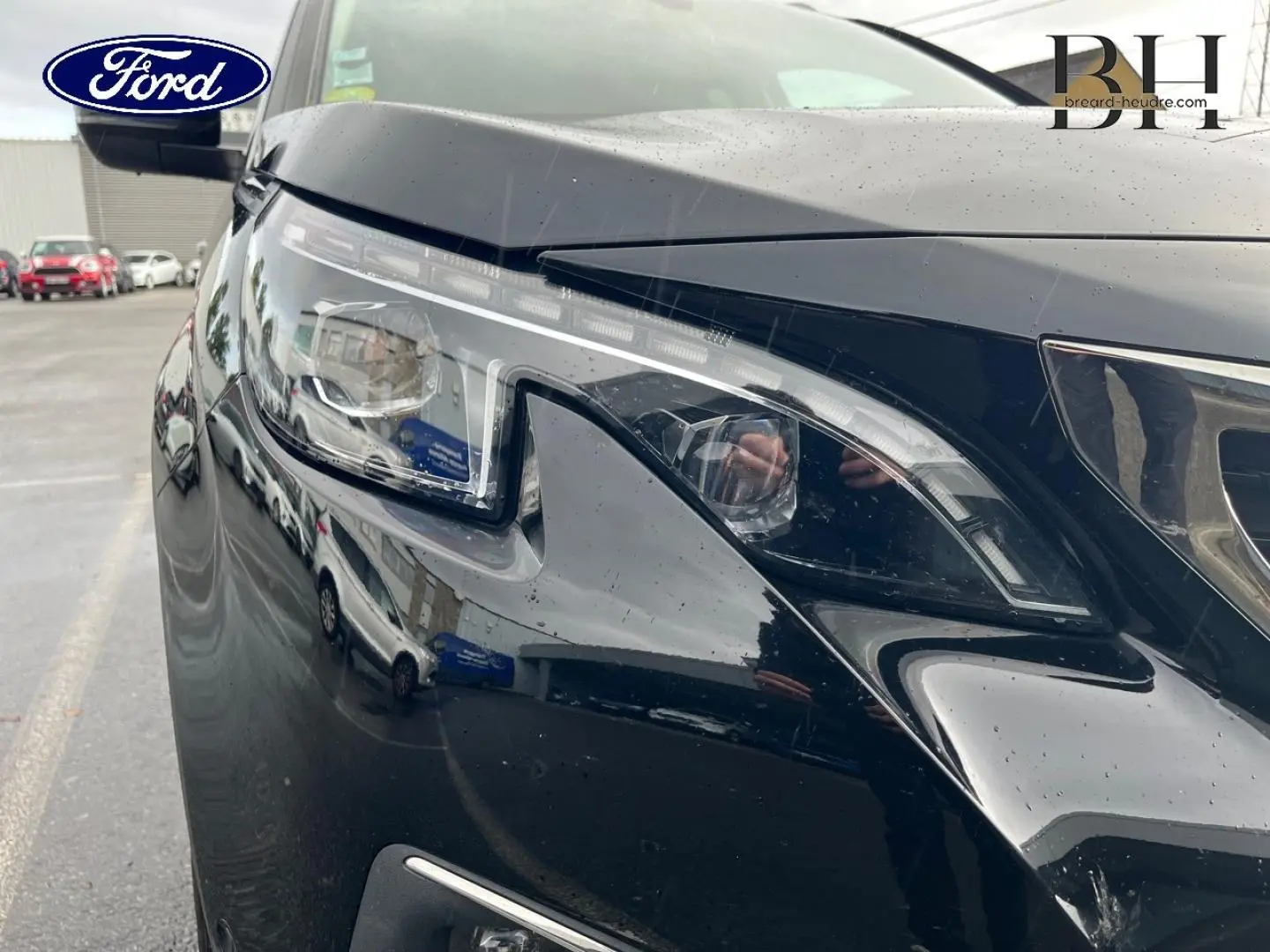 Gros plan sur le phare avant droit noir Perla Nera du Peugeot 5008 avec reflets et pluie sur la carrosserie.