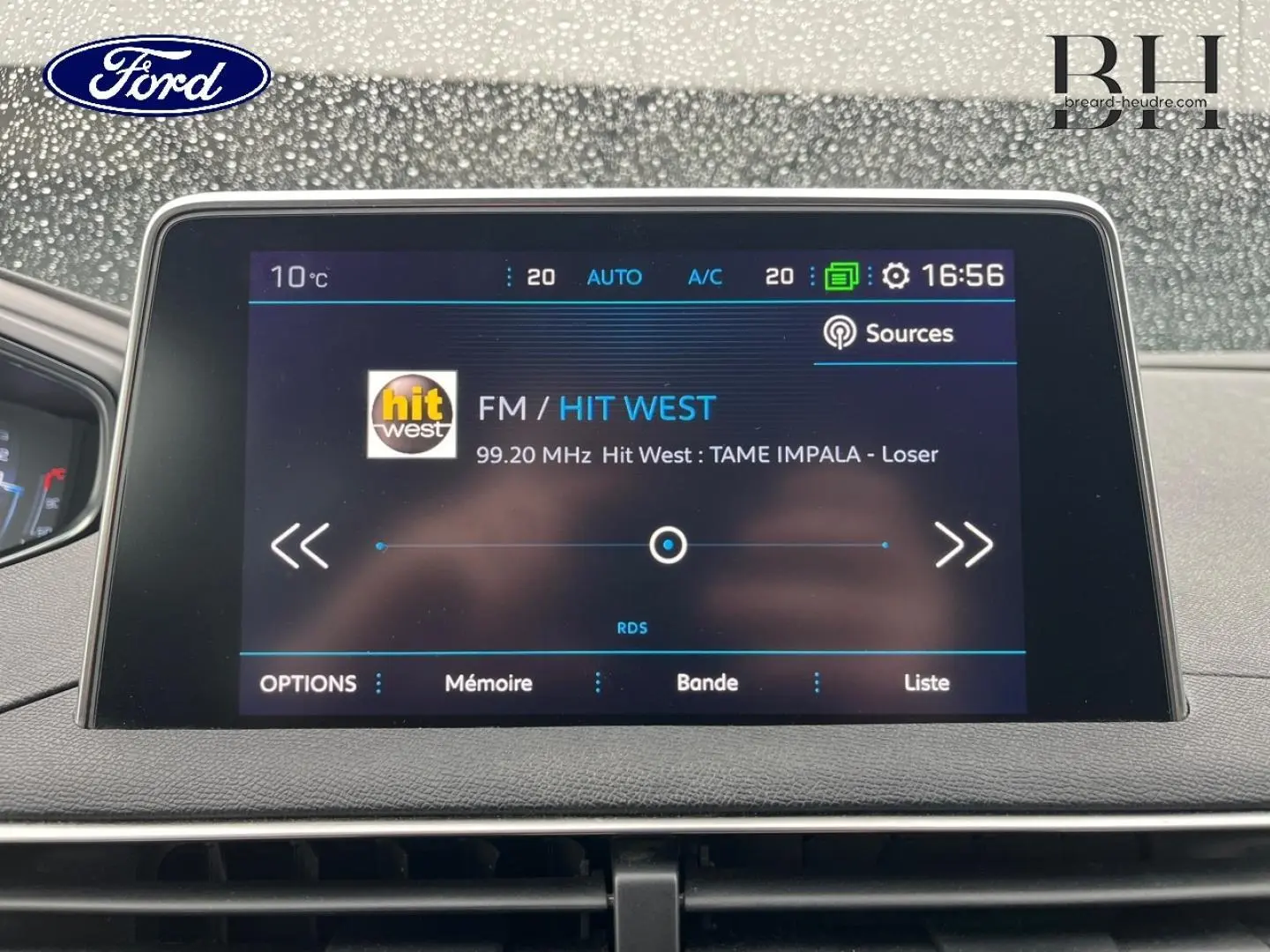 Écran tactile central d'infodivertissement Peugeot 5008 noir, affichant la radio FM Hit West à 16h56 par temps pluvieux.
