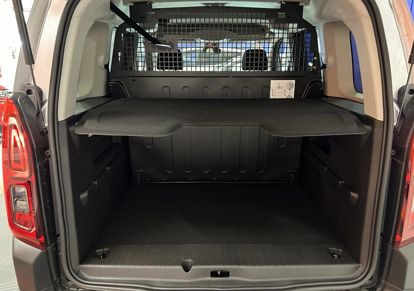 Coffre ouvert vu de l'arrière d'un Citroën Berlingo Multispace gris avec séparation grillagée et tablette de rangement.