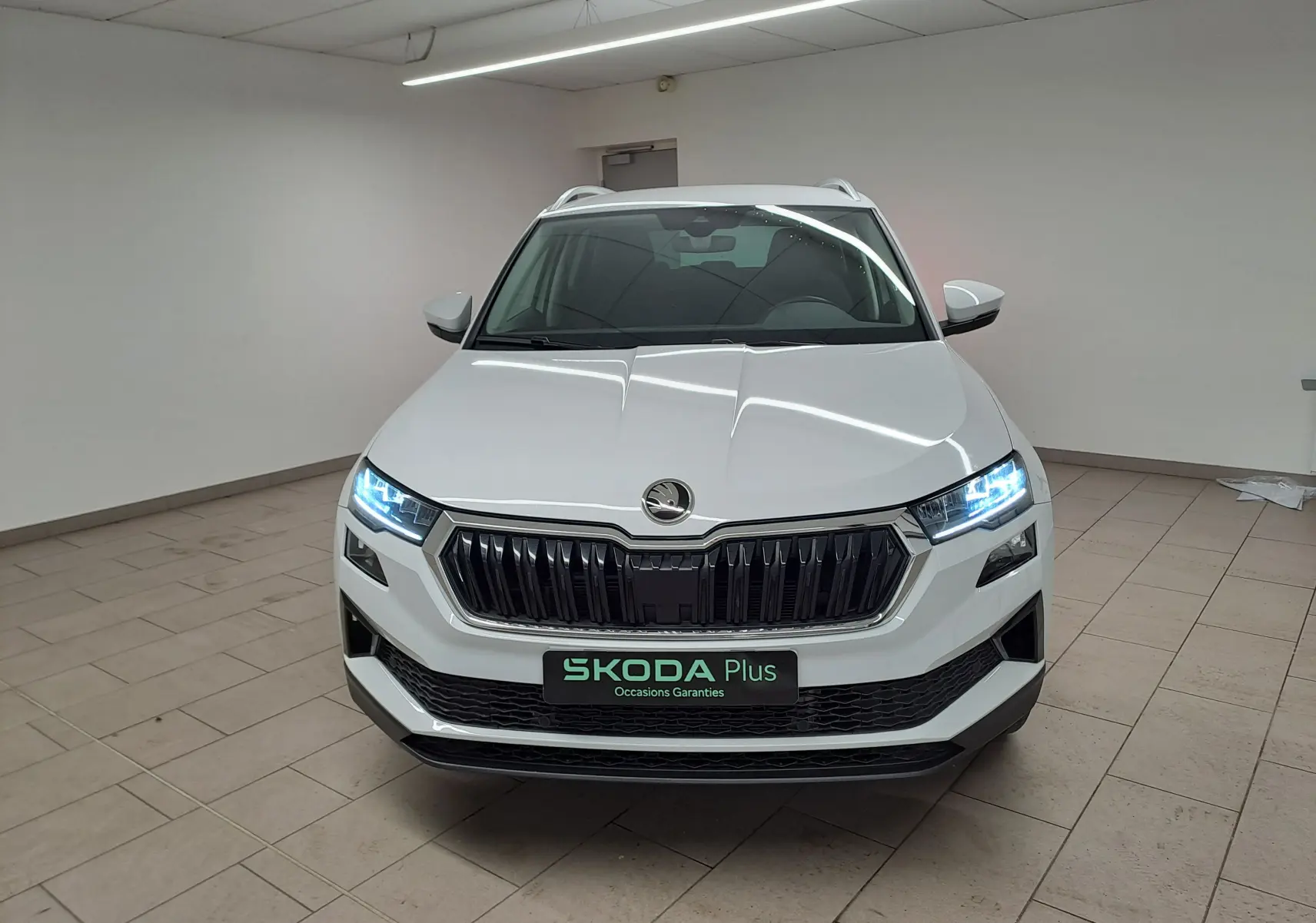 Vue avant d’un Skoda Karoq blanc Lune 2025 avec calandre noire, phares allumés et pack chrome extérieur visible.