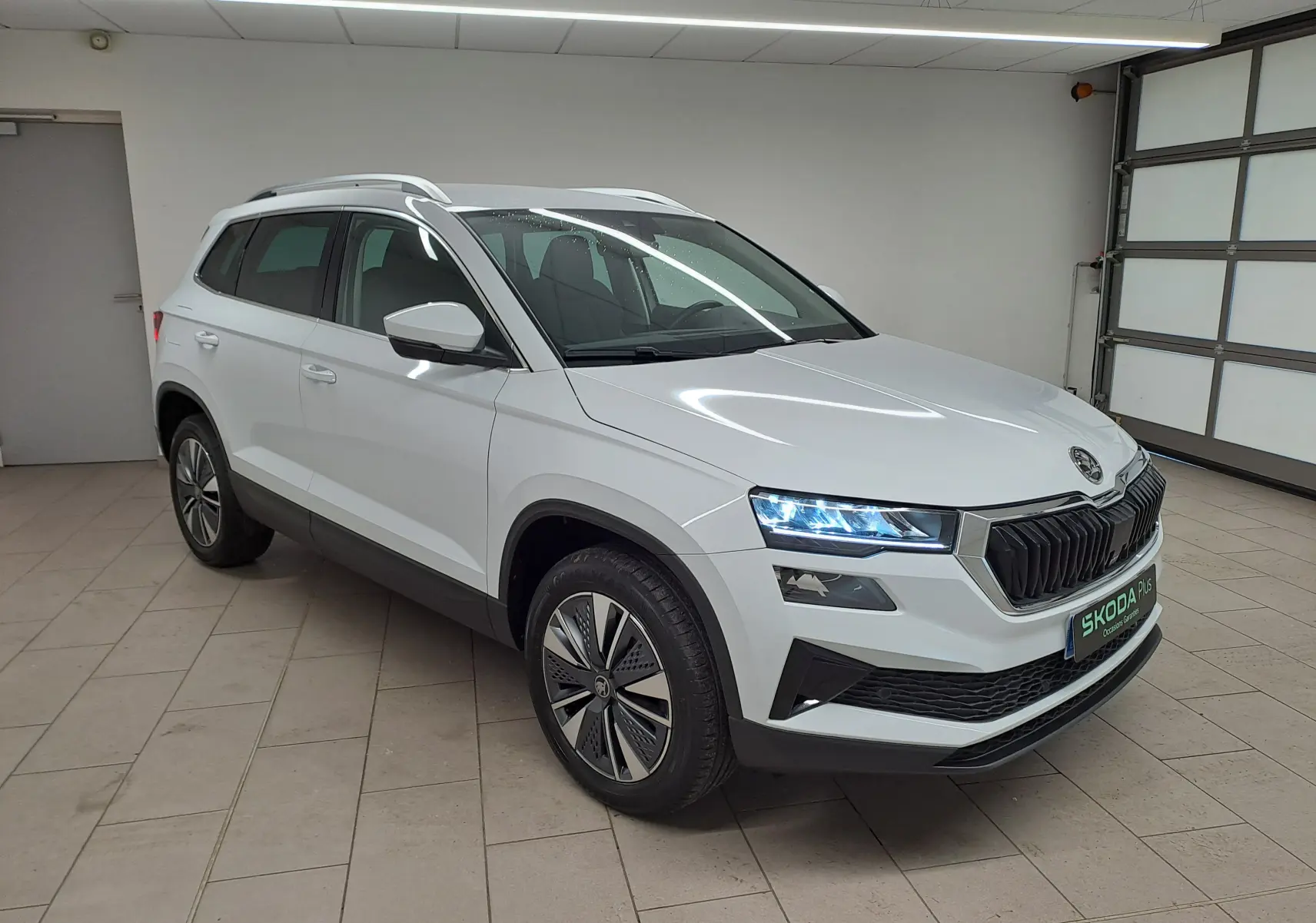 Vue 3/4 avant droite du Skoda Karoq blanc Lune avec calandre noire, jantes spécifiques et rampes de pavillon argentées.