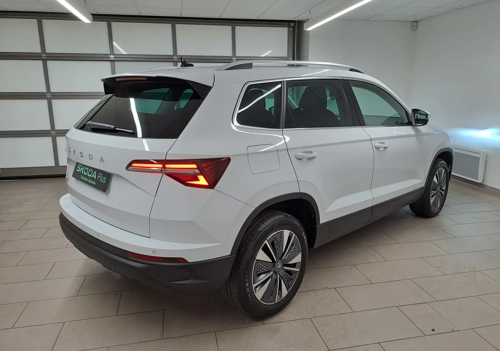 Vue 3/4 arrière droite du Skoda Karoq blanc Lune, avec feux arrière allumés et détails chromés autour des vitres.