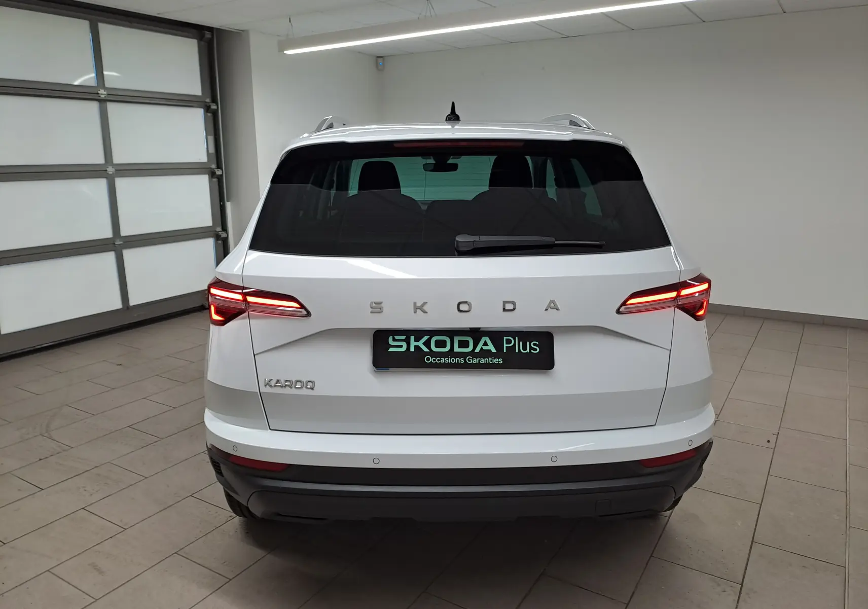 Vue arrière d'un Skoda Karoq blanc Lune 2025 avec feux allumés et détails chromés sur un sol carrelé en intérieur.