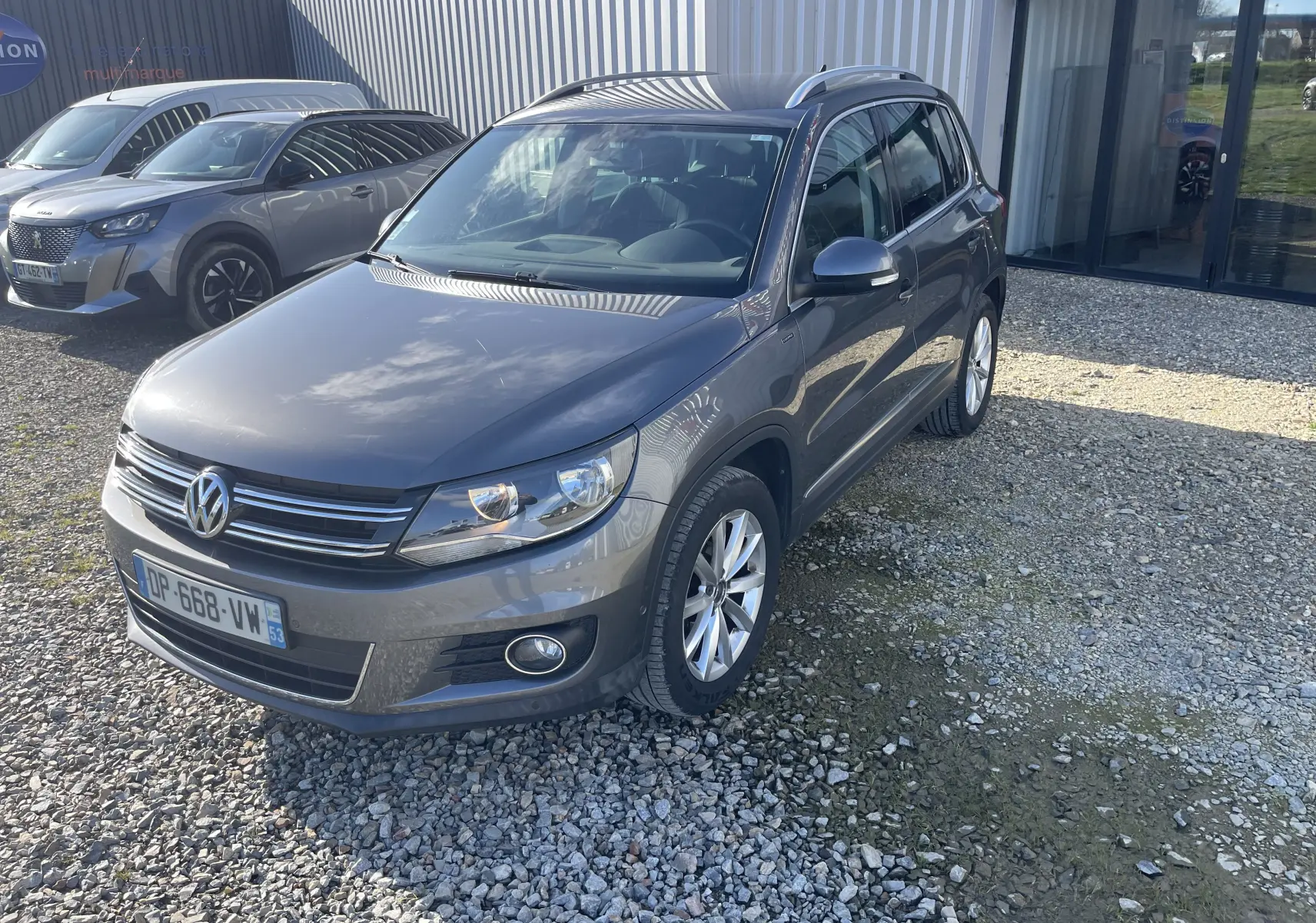 Volkswagen Tiguan gris foncé en 3/4 avant droit, avec jantes alliage et calandre chromée visible, stationné sur gravier.