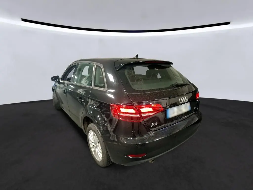 Audi A3 Sportback noire vue 3/4 arrière droit, avec feux arrière allumés et becquet sur le toit.