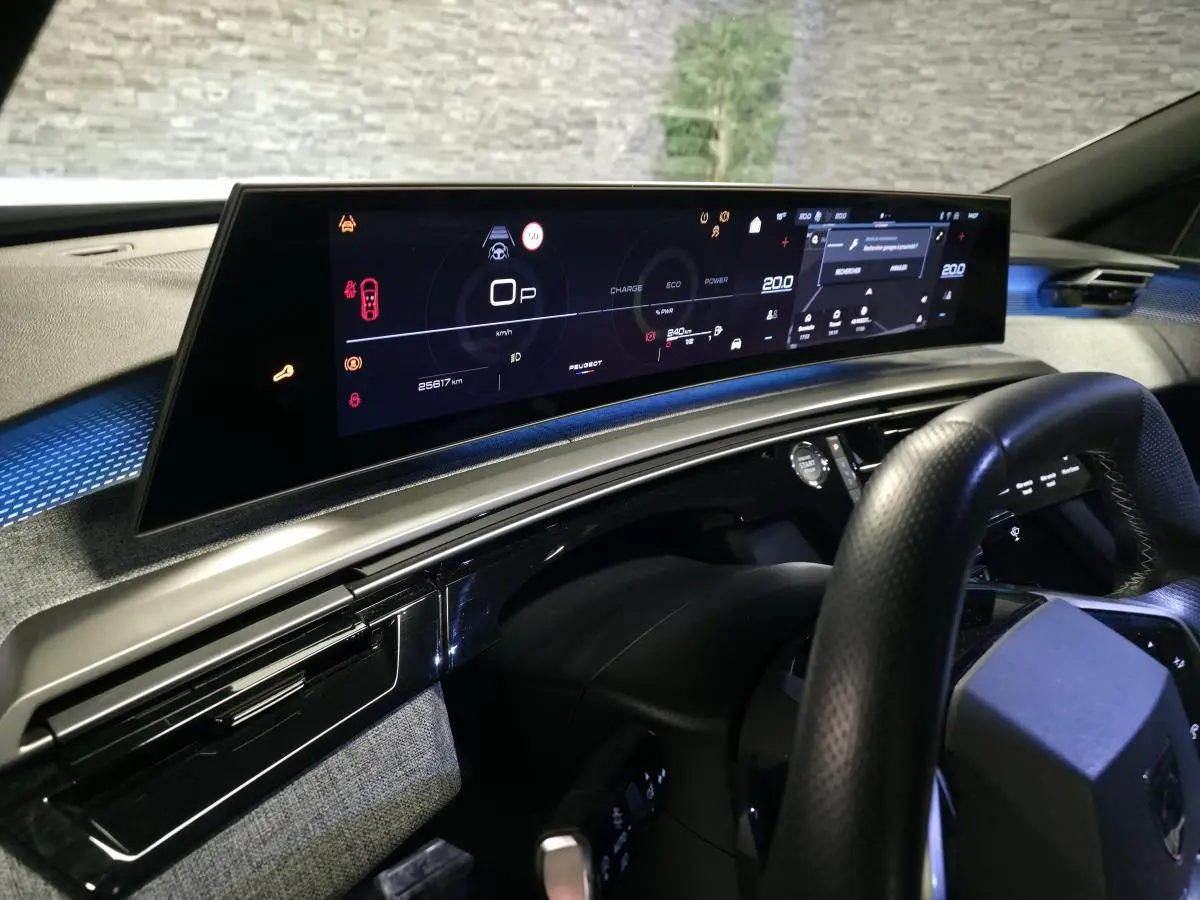 Vue intérieure du tableau de bord numérique panoramique du Peugeot 3008 hybride 2025, avec volant noir et finitions tissu gris.