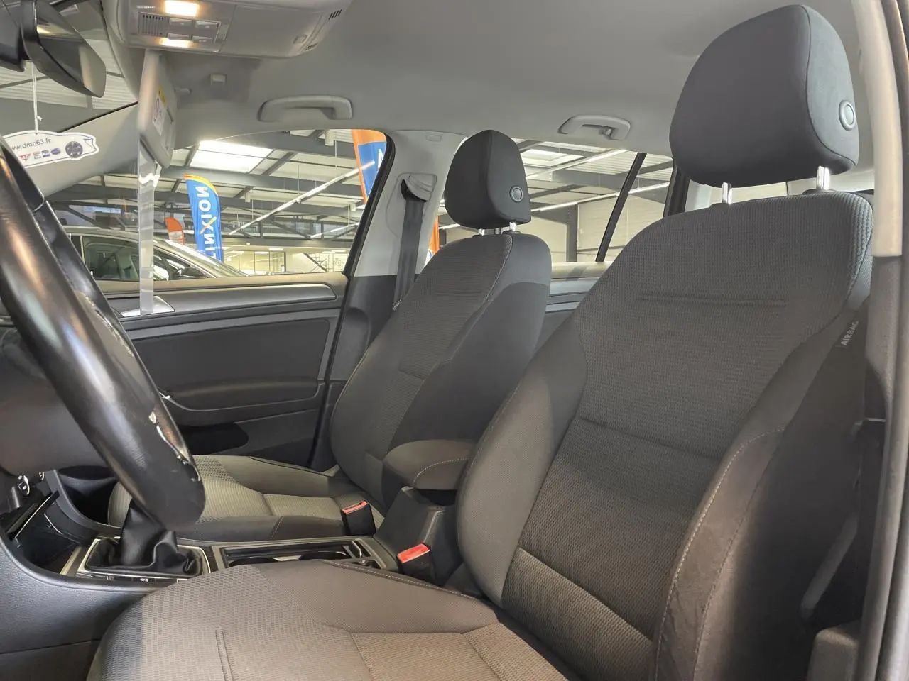 Intérieur avant de la Volkswagen Golf SW 2017, sièges tissu gris foncé avec accoudoir central et volant cuir.