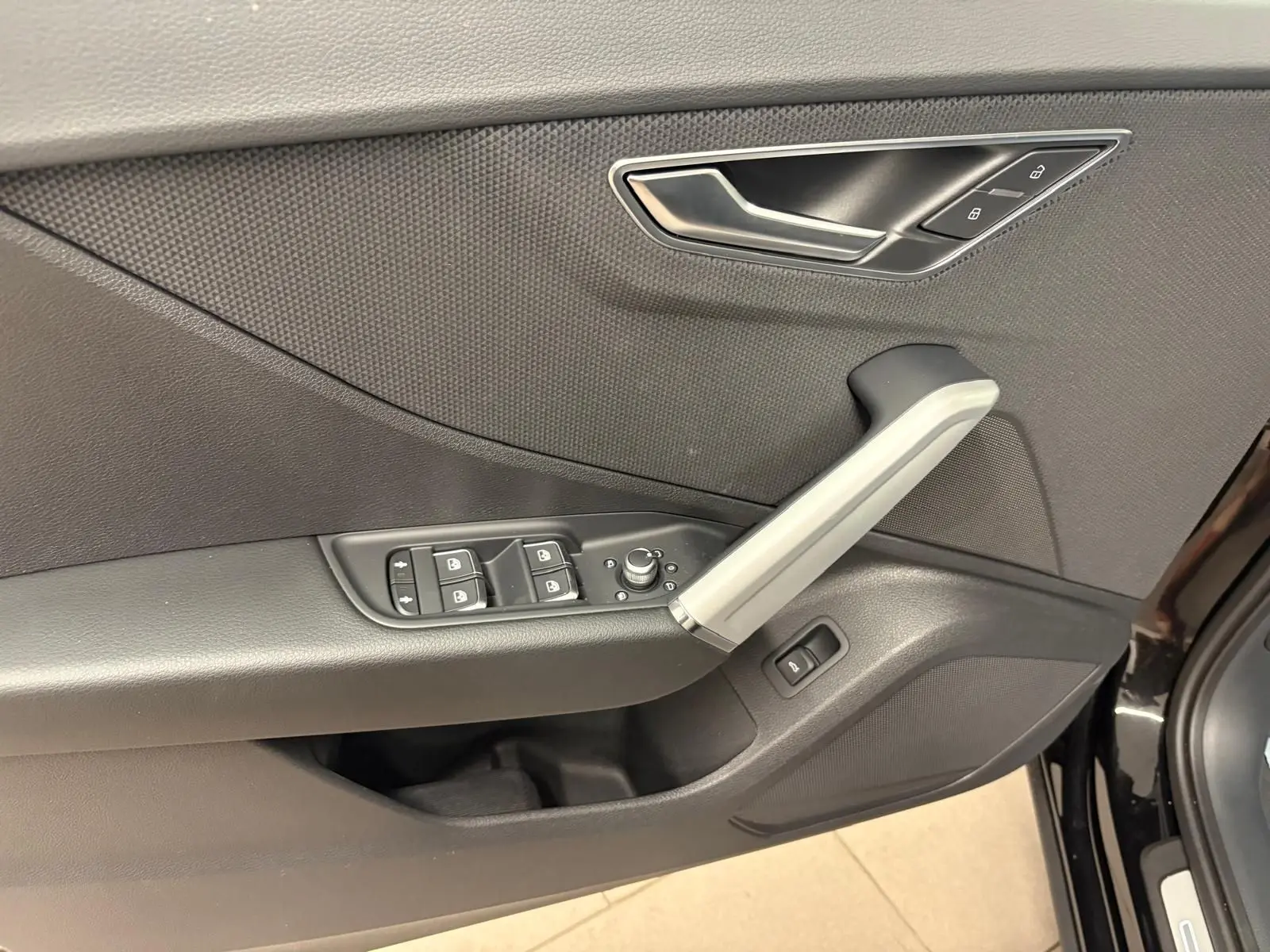 Gros plan sur la porte avant gauche de l'Audi Q2 Noir Mythic, montrant les commandes des vitres et la poignée intérieure argentée.
