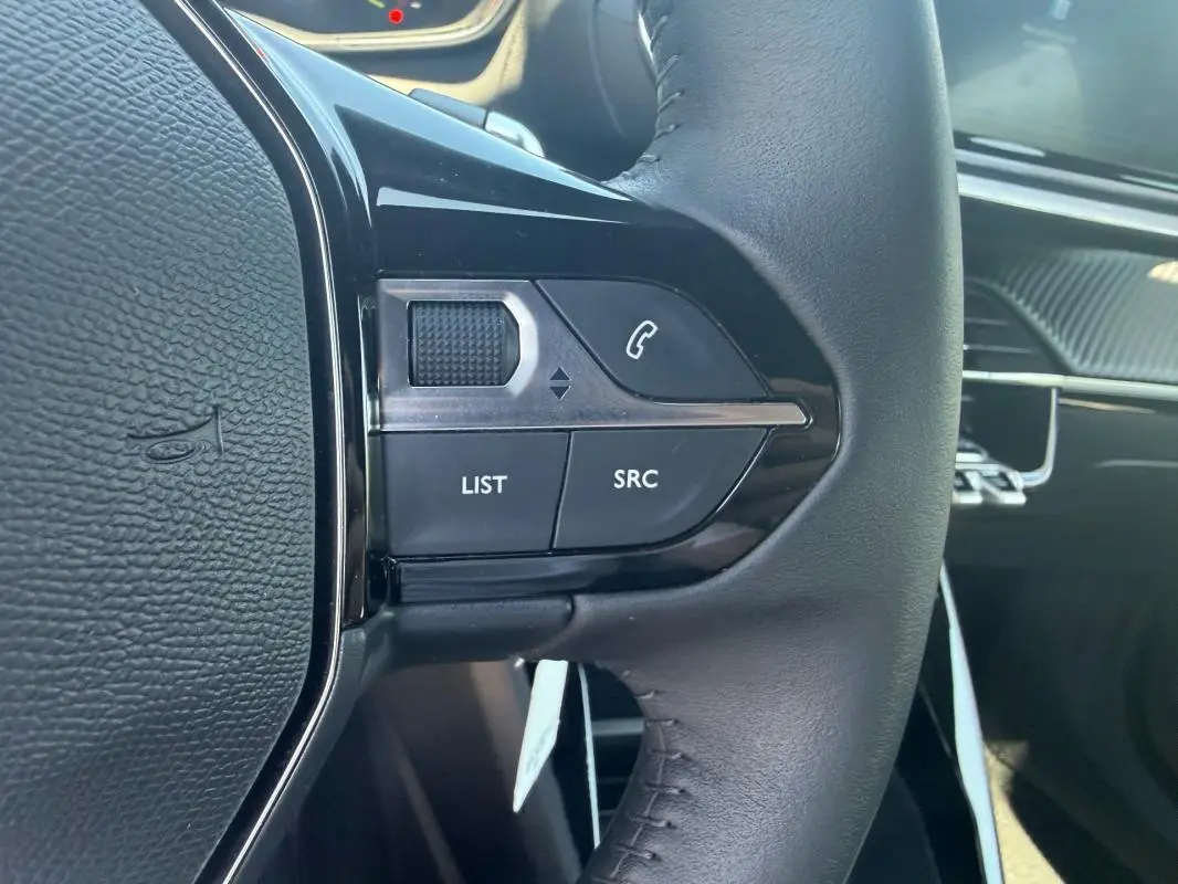 Gros plan sur les commandes gauche du volant noir du Peugeot 2008 Hybrid 136 e-DCS6 Style, avec tableau de bord en arrière-plan.