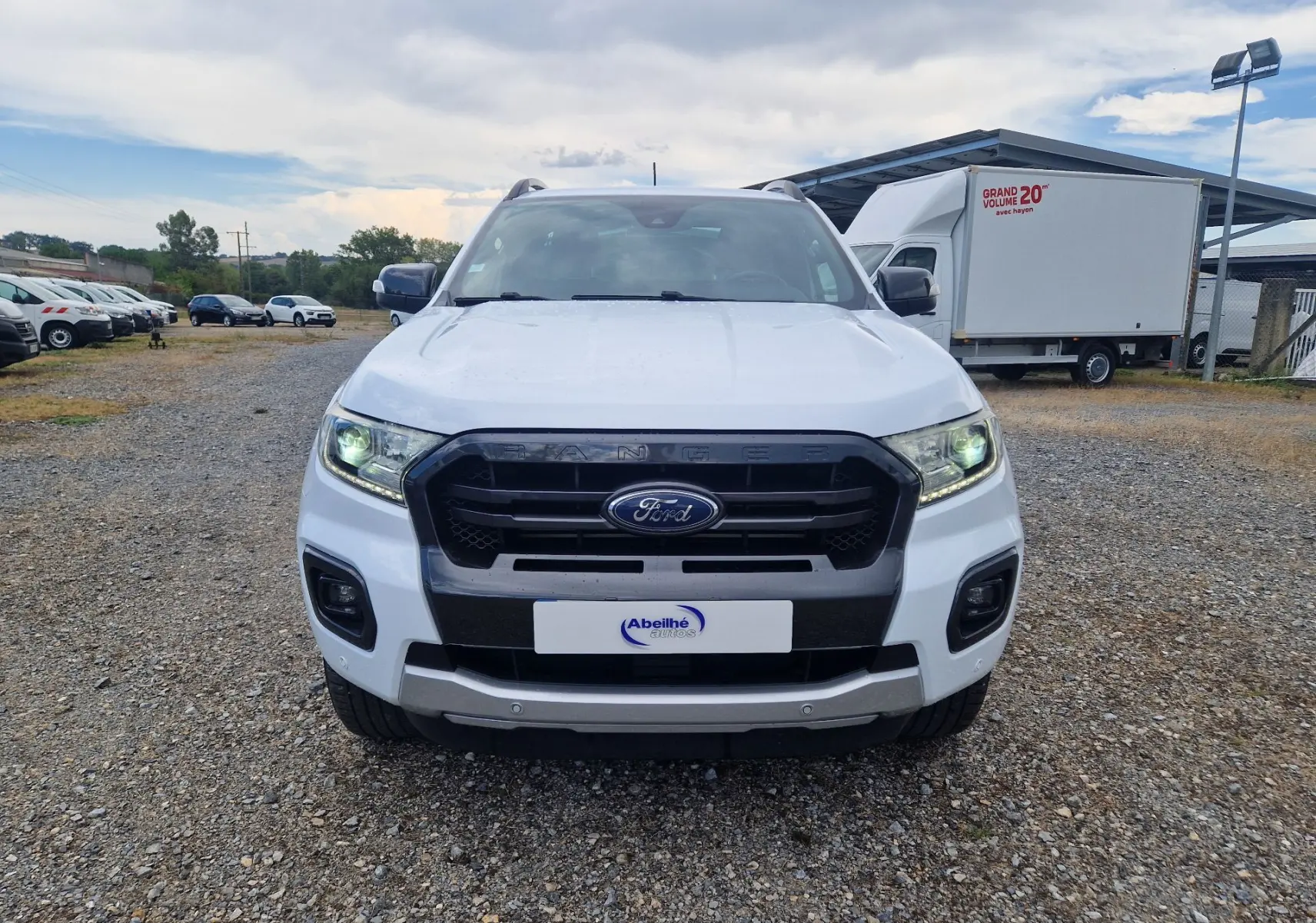 Vue avant d'un Ford Ranger Super Cabine blanc 4x4 avec calandre noire et phares allumés sur un terrain extérieur.