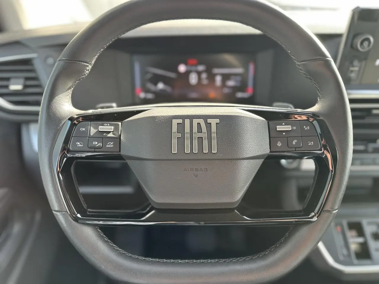 Vue rapprochée du volant noir FIAT avec commandes intégrées et tableau de bord numérique flou en arrière-plan.