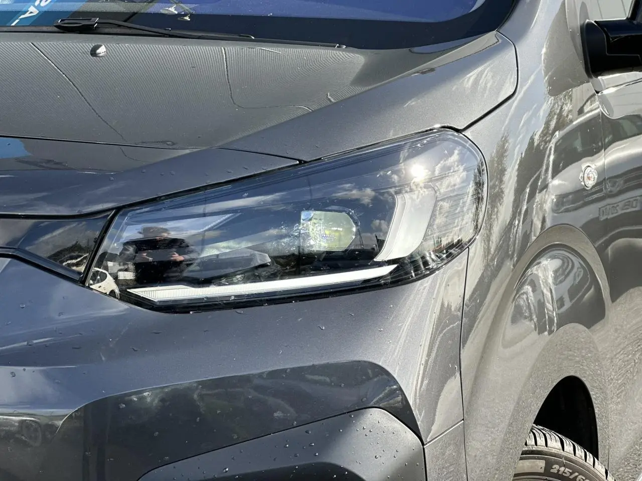 Gros plan sur le phare avant droit du FIAT Scudo Fourgon gris Platinium avec carrosserie lisse et reflets lumineux.
