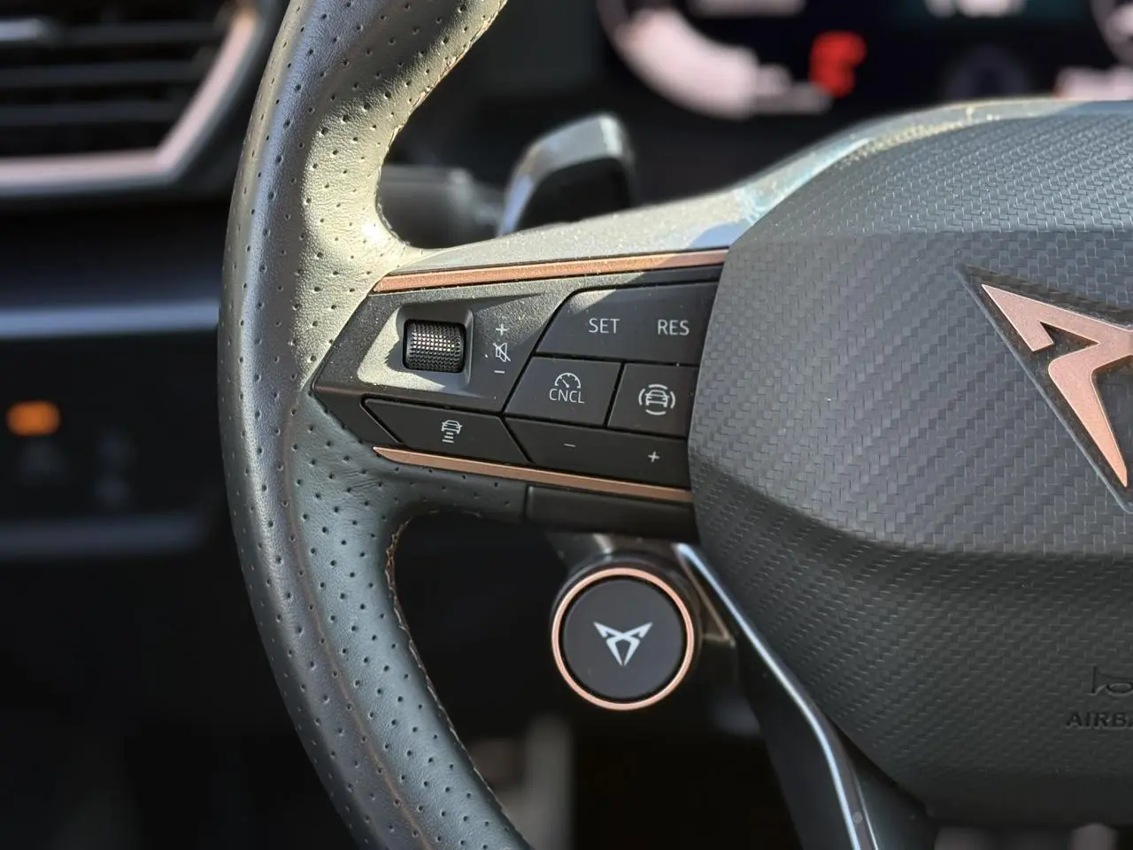 Gros plan sur le volant perforé noir du CUPRA LEON ST avec commandes intégrées et logo cuivre au centre.