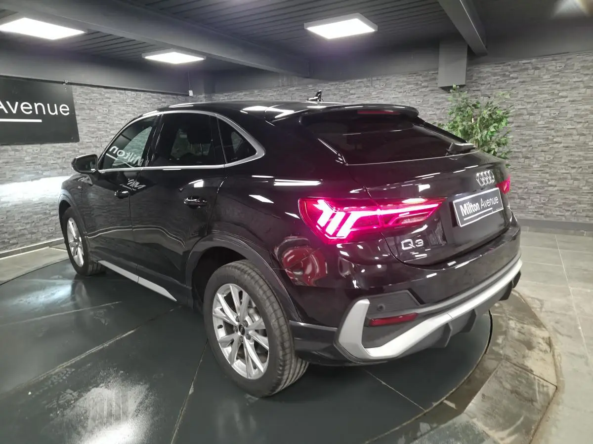 Audi Q3 Sportback noir vue 3/4 arrière droit, feux arrière allumés et vitres surteintées en intérieur showroom.
