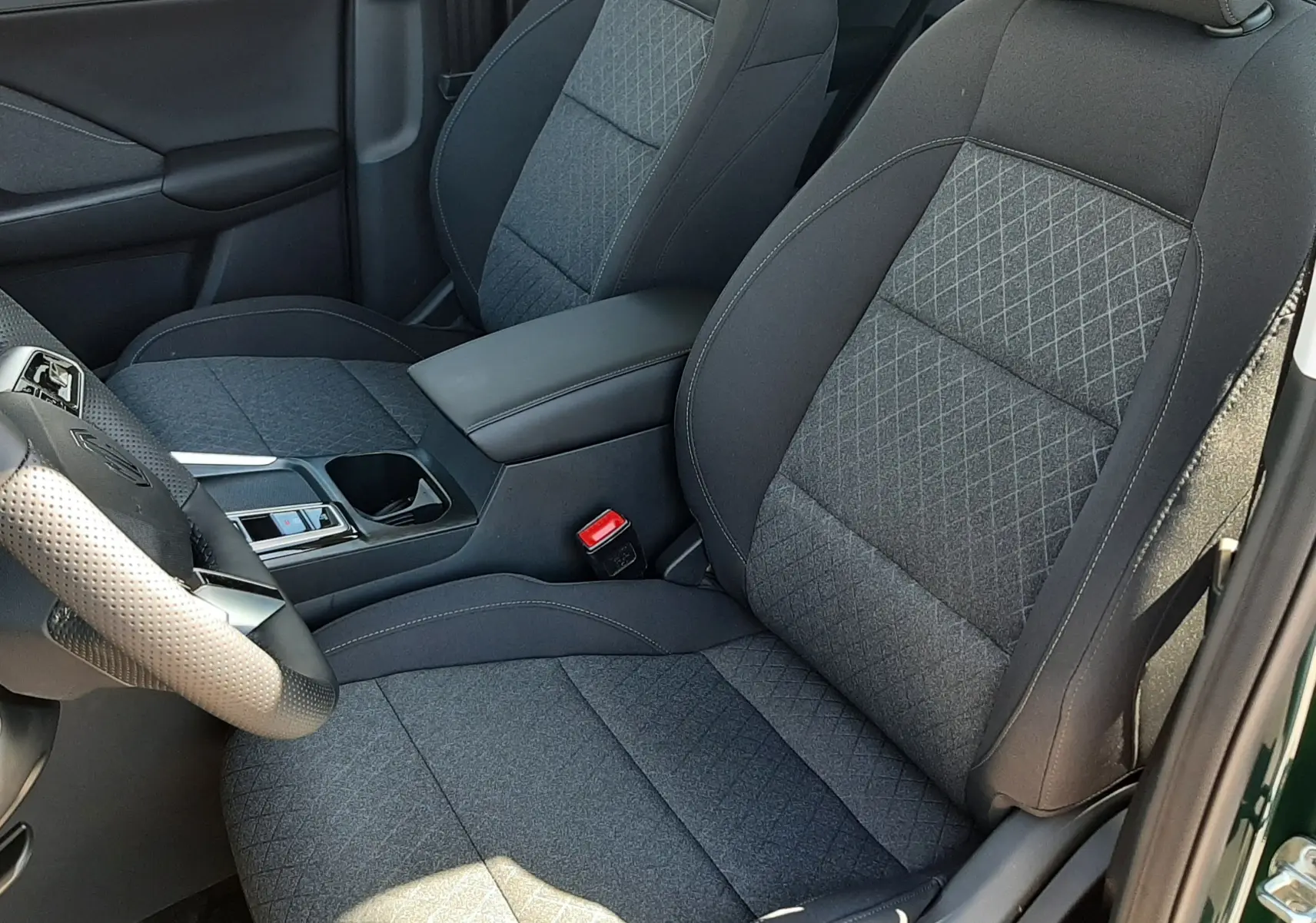 Vue intérieure côté conducteur du MG ZS 1.5 Hybrid+ 2026, sièges tissu gris avec surpiqûres losanges et accoudoir central.