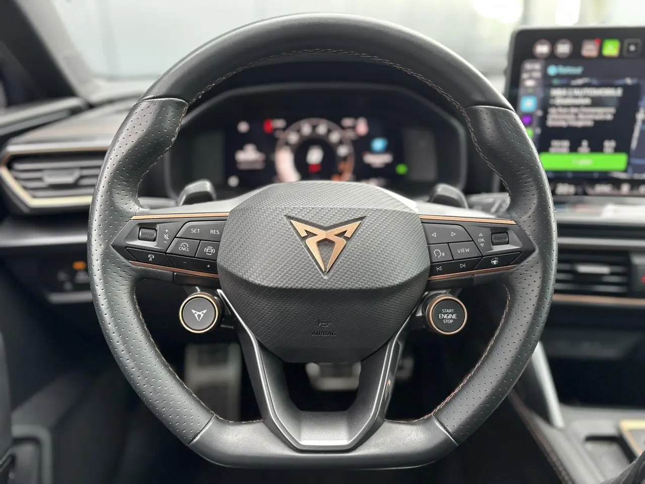 Volant cuir perforé noir avec logo CUPRA cuivré et commandes intégrées, tableau de bord numérique en arrière-plan.