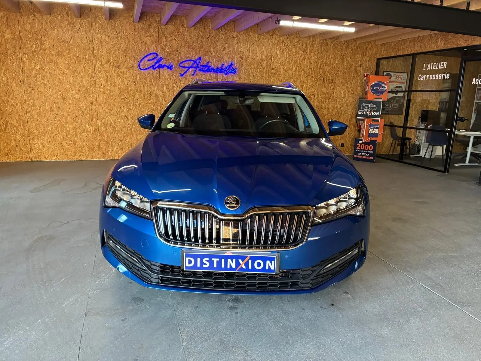Vue de face d'une Skoda Superb Combi bleu métallisé, avec calandre chromée et phares LED dans un showroom.