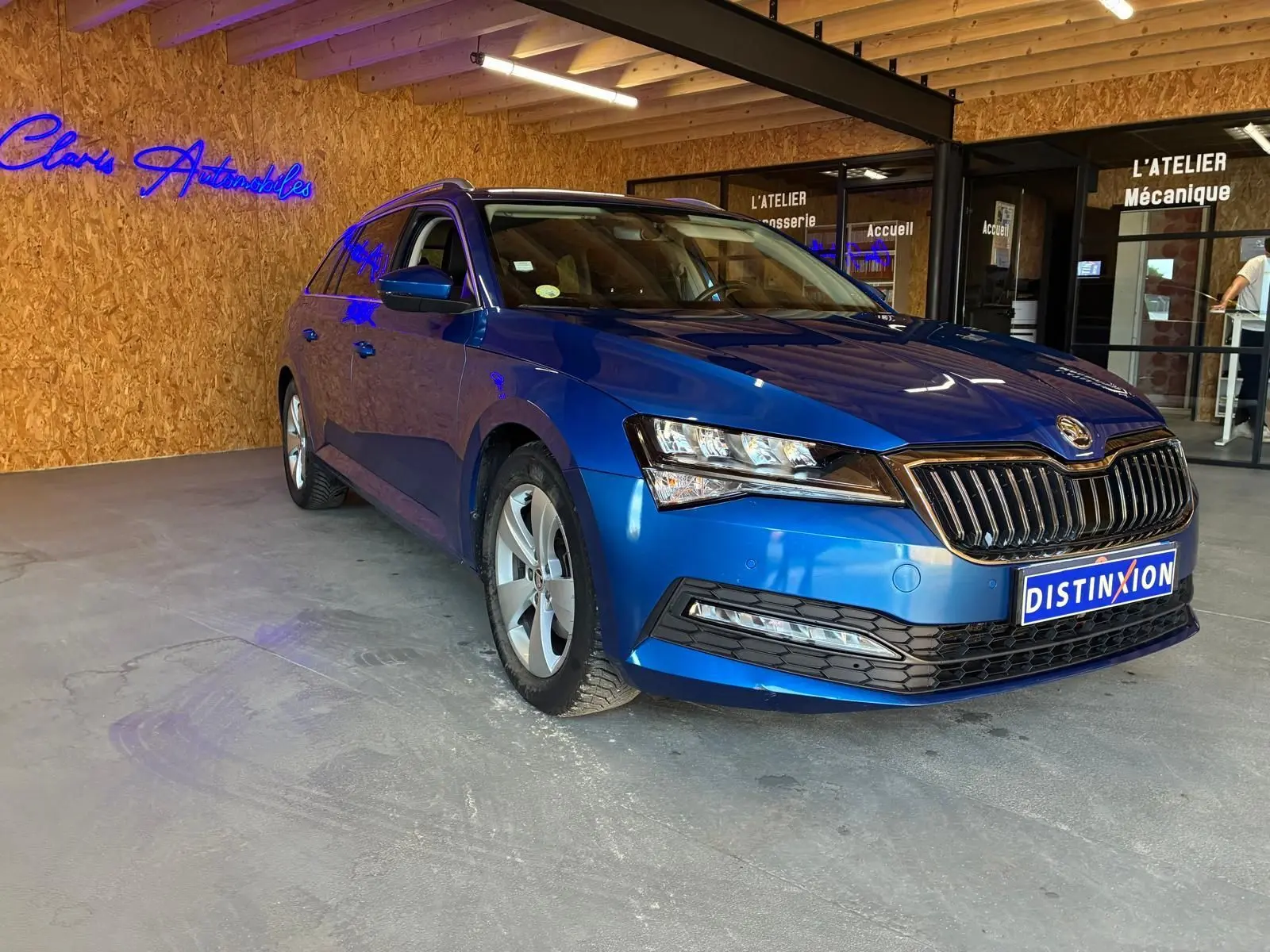 Skoda Superb Combi bleu en 3/4 avant droit, stationné en intérieur devant un mur en bois avec enseigne néon bleue.