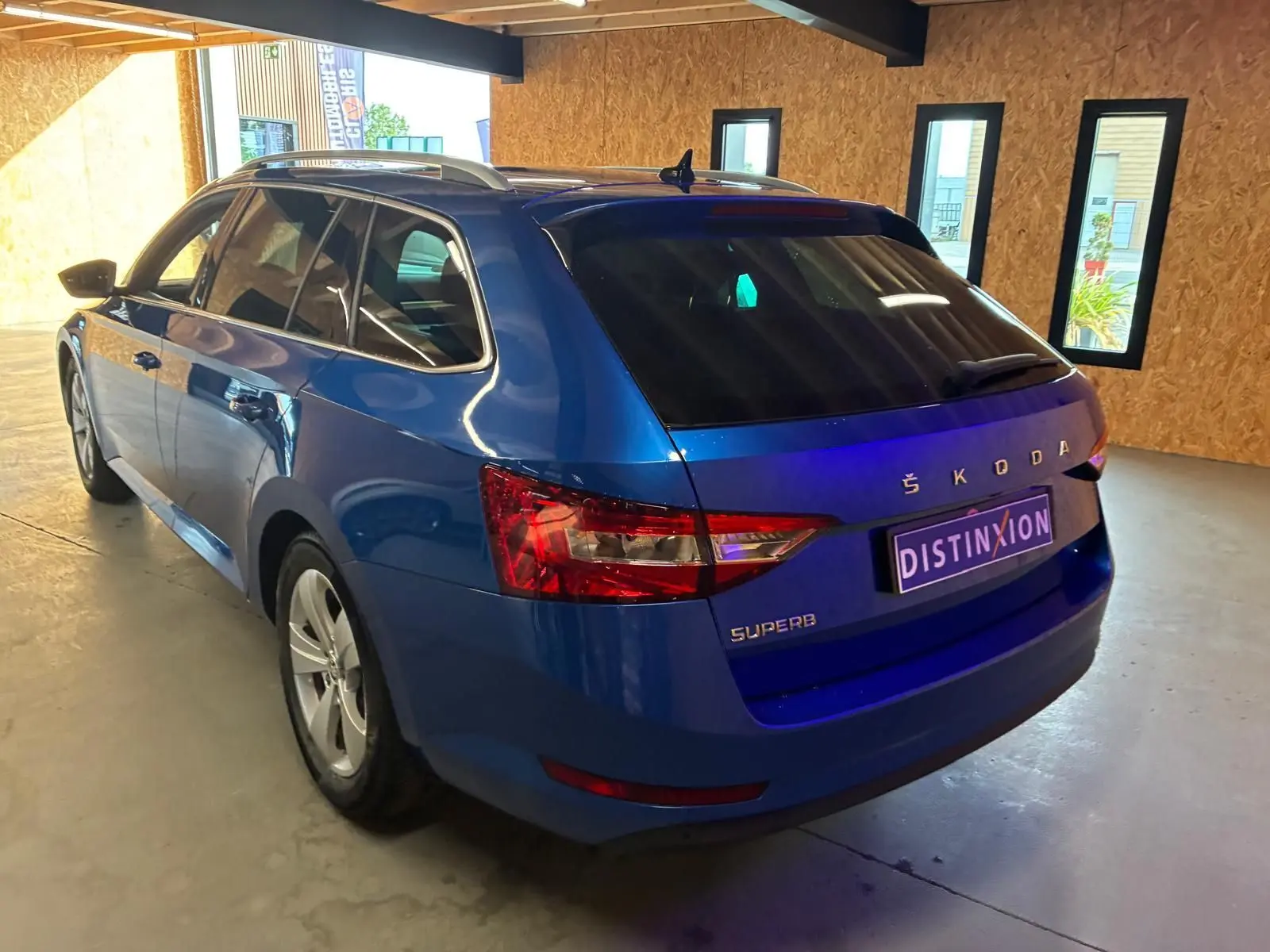 Vue 3/4 arrière gauche d'un break Skoda Superb Combi bleu dans un intérieur avec éclairage naturel.