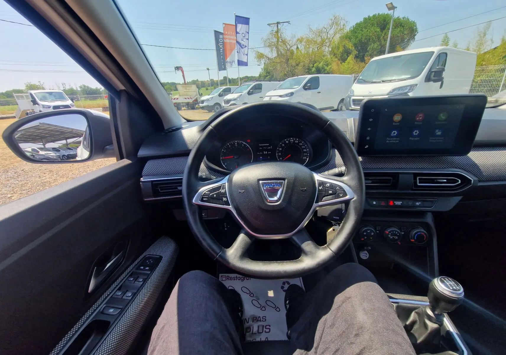 Vue intérieure centrée sur le volant Dacia Sandero blanc, tableau de bord et levier de vitesses manuelle visibles.