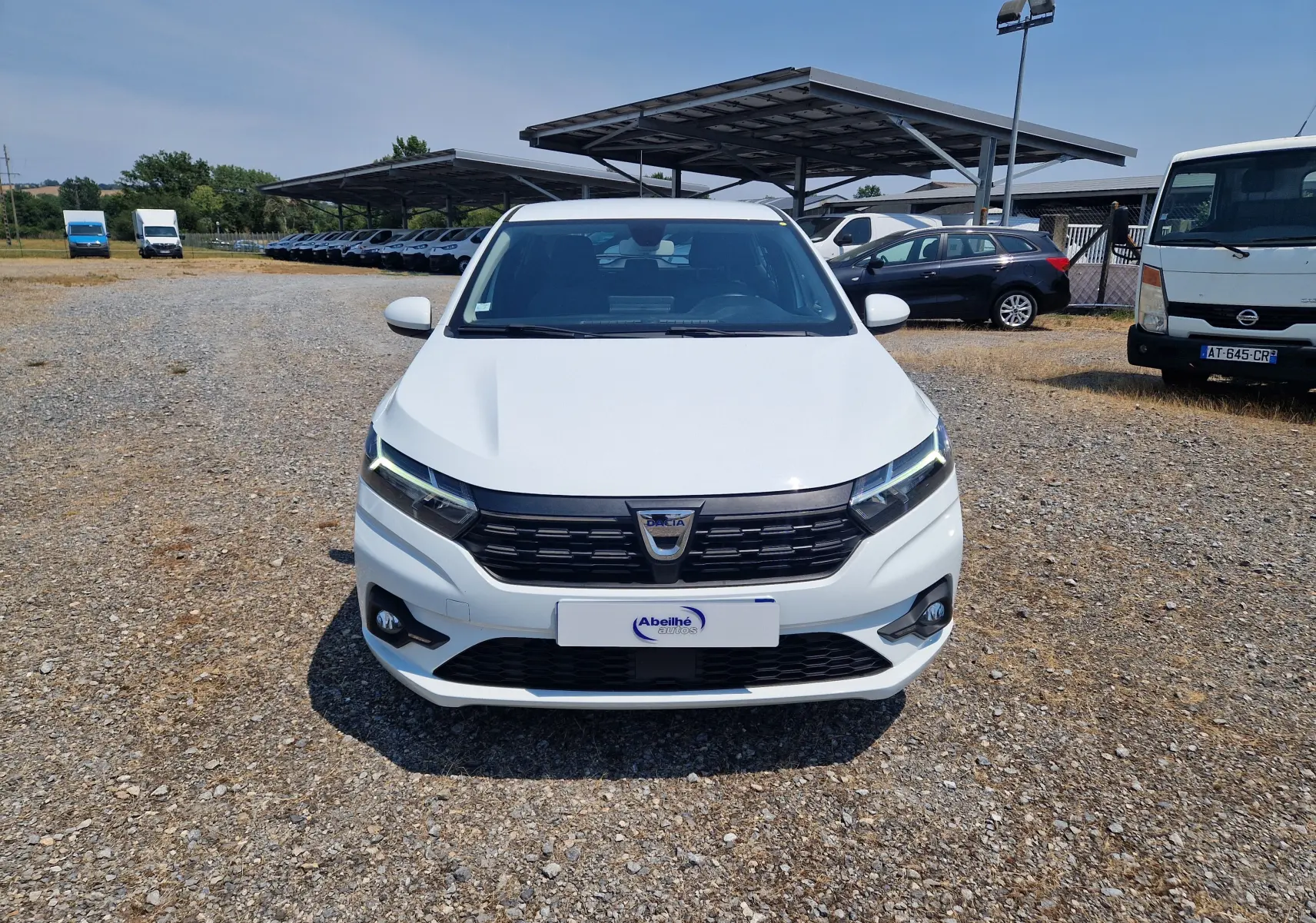Vue frontale d'une Dacia Sandero blanche 2022 avec calandre noire et feux LED allumés sur un parking extérieur.
