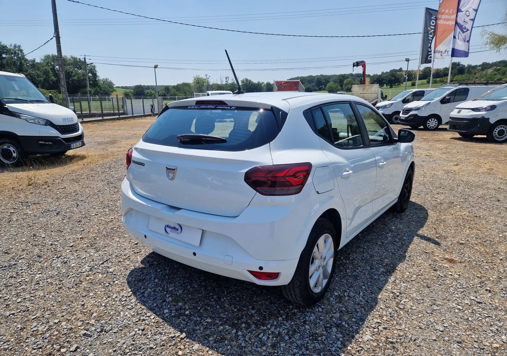 Vue 3/4 arrière droite d'une Dacia Sandero blanche 2022, version Confort, sur un terrain gravillonné en extérieur.