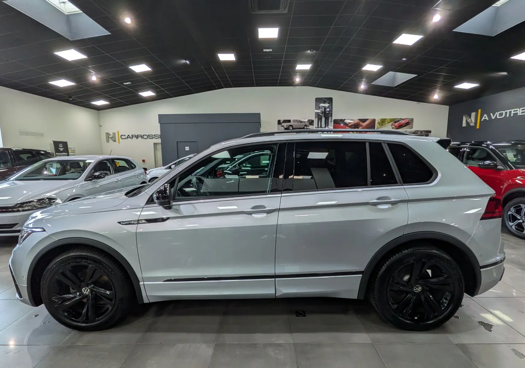 Vue de profil côté gauche d’un Volkswagen Tiguan 2024 reflet d’argent avec jantes noires et vitres teintées en showroom.