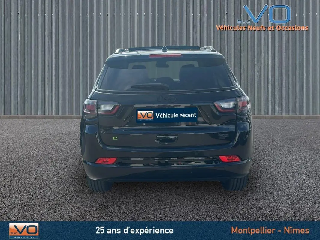 Vue arrière d'un Jeep Compass noir 2023 avec barres de toit et feux arrière LED allumés sur fond industriel.