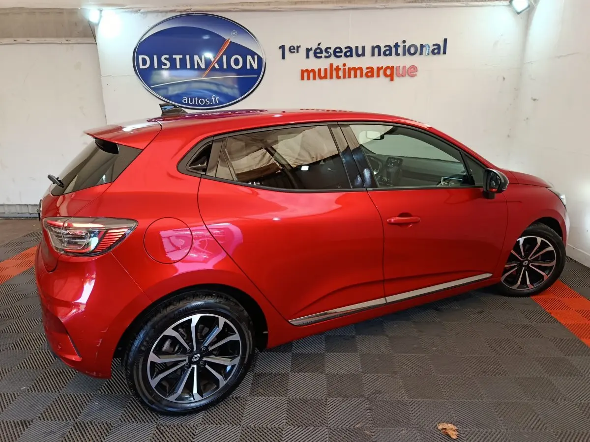 Profil droit d'une Renault Clio Techno TCe 90 rouge 2024 avec jantes alliage et vitres teintées en intérieur showroom.