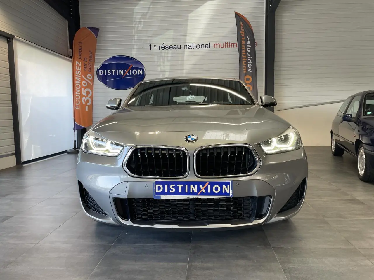 Vue frontale d'une BMW X2 M Sport X gris foncé avec phares LED allumés en intérieur showroom.