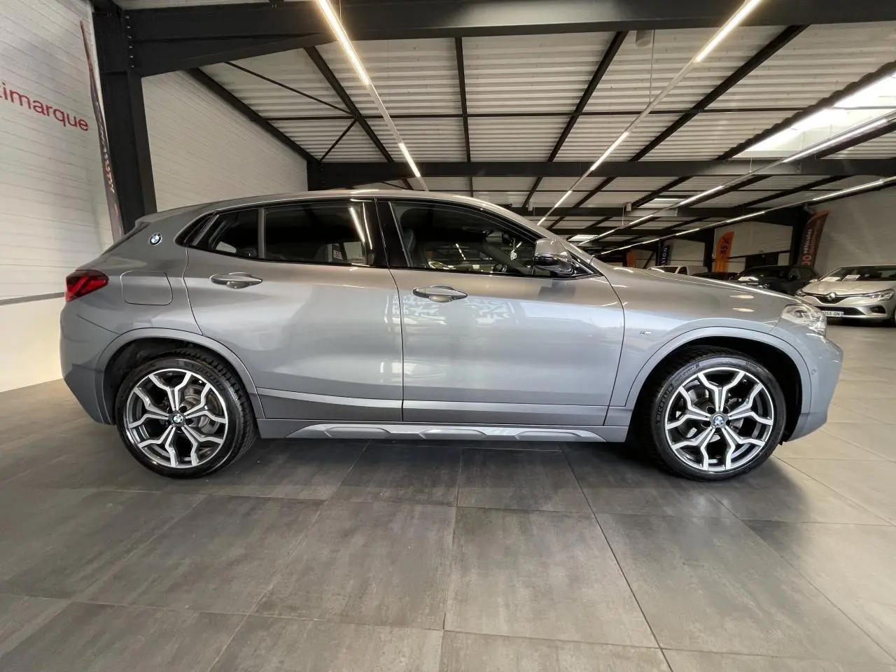 BMW X2 sDrive 18d gris foncé vue de profil côté gauche avec jantes alliage 19 pouces style 722 M en intérieur showroom.