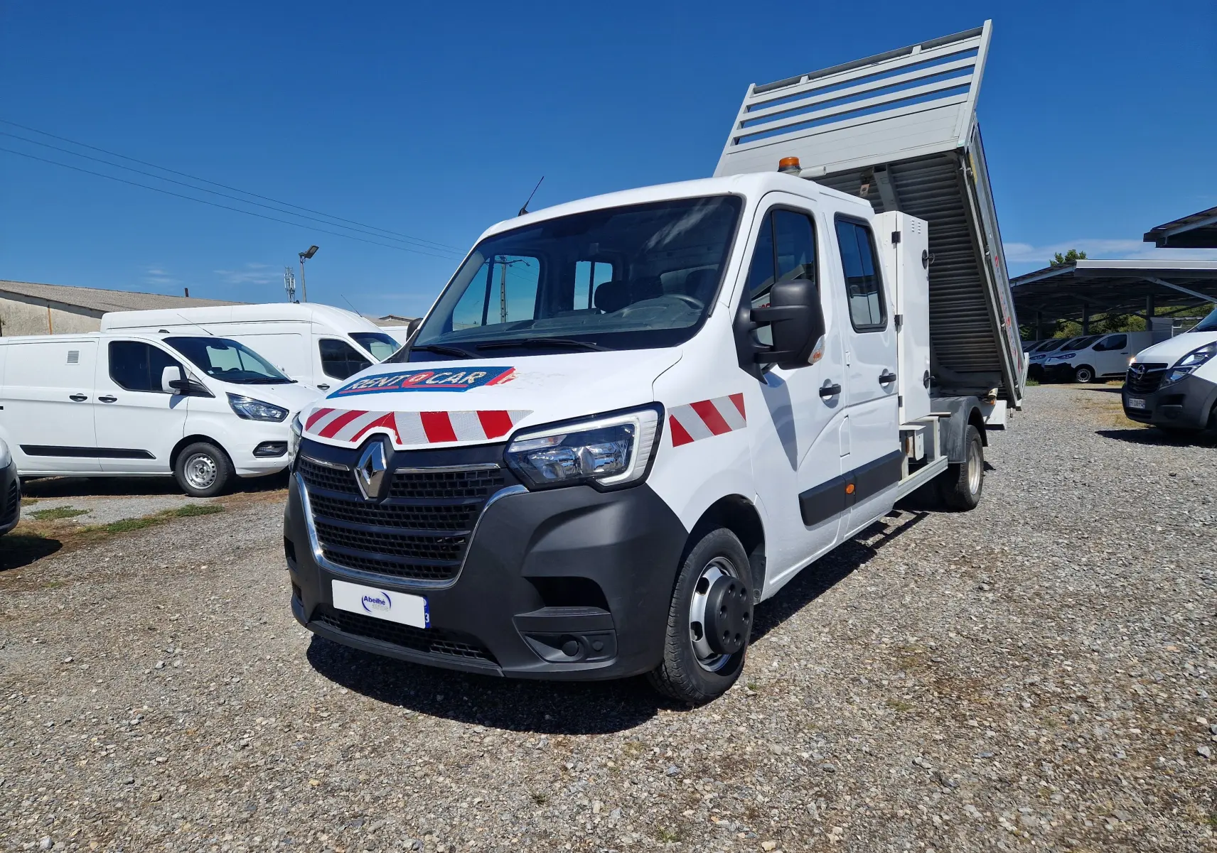 Renault Master blanc en 3/4 avant droit avec benne relevée et marquages rouges et blancs sur la carrosserie.